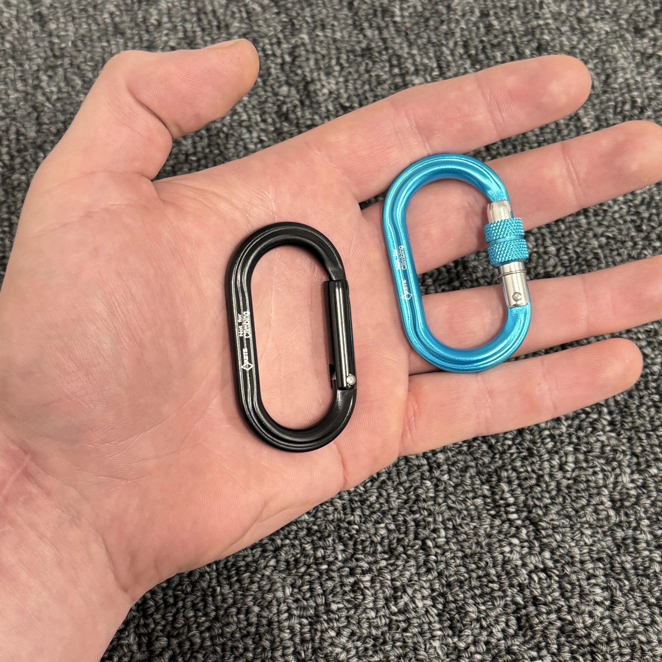 Hand holding two carabiners, one black and one blue, against a textured gray background